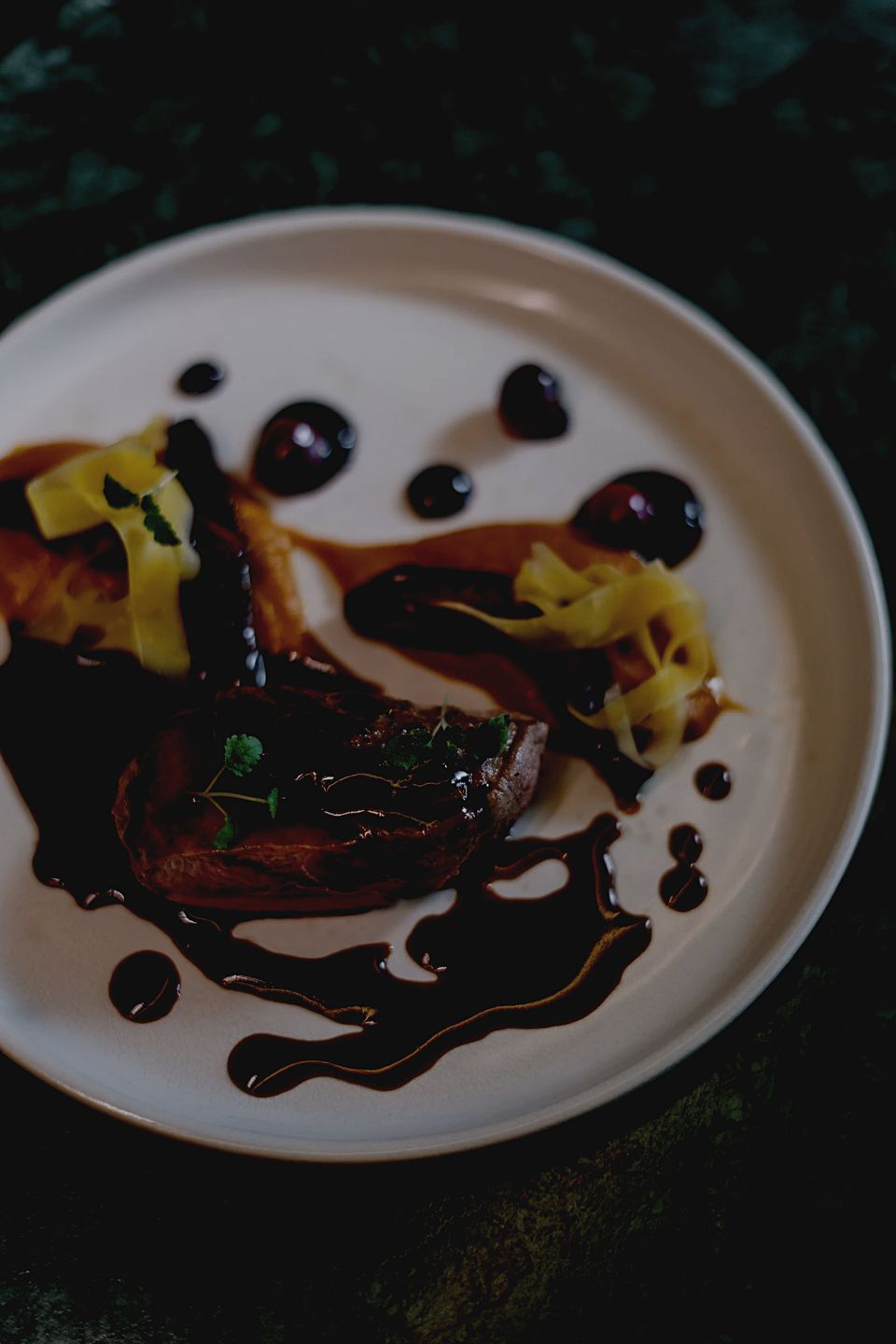 A plated dish of seared meat with glossy dark sauce droplets, pale yellow accents and fresh micro-herbs on a white plate