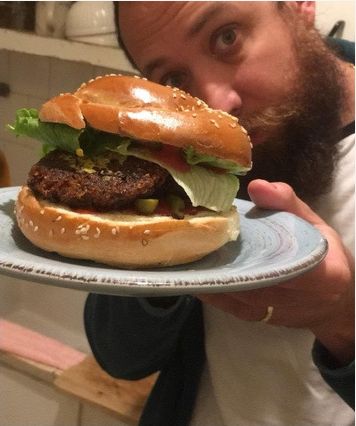 Felix Kris peering over a large sesame-bun burger with a thick patty, lettuce, tomato and pickles on a light blue plate