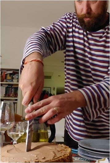 Felix Kris in a blue and white striped shirt slicing a large round cake with a kitchen knife at a dining table