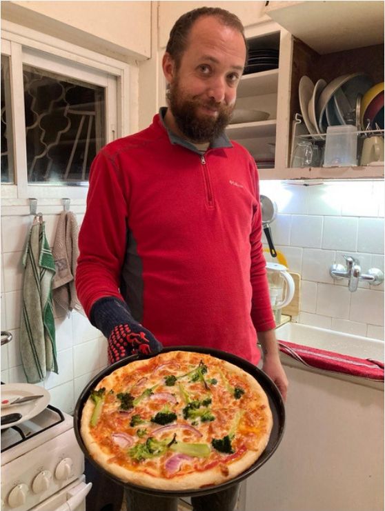 Felix Kris in a red fleece holding a round pizza pan with a golden-crust pizza topped with broccoli and red onion