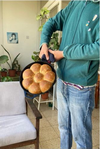 Felix Kris in a teal hoodie and oven mitt holding a black cast-iron skillet of golden pull-apart bread topped with sesame seeds