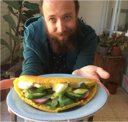 Felix Kris smiling and holding out a light blue plate with a folded yellow flatbread filled with greens, sliced cucumber, radish and sliced egg