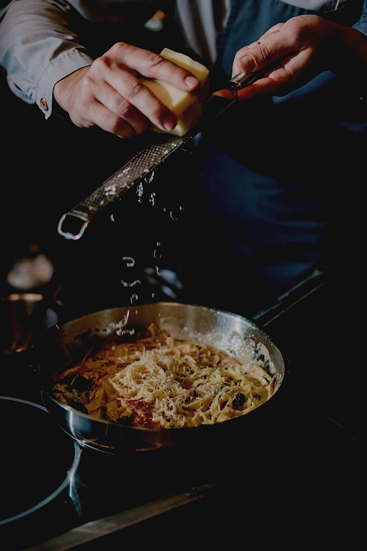 Felix Kris grating fresh cheese over a pan of creamy flat pasta with broccoli, with the grated cheese falling onto the dish