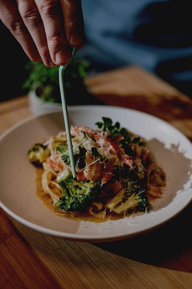 A hand using light green tweezers to garnish a plate of flat pasta with broccoli, sun-dried tomato, grated cheese and microgreens