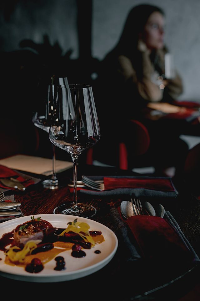 A plated dish of glazed meat with orange purée and dark berries beside empty wine glasses on a dark wooden table