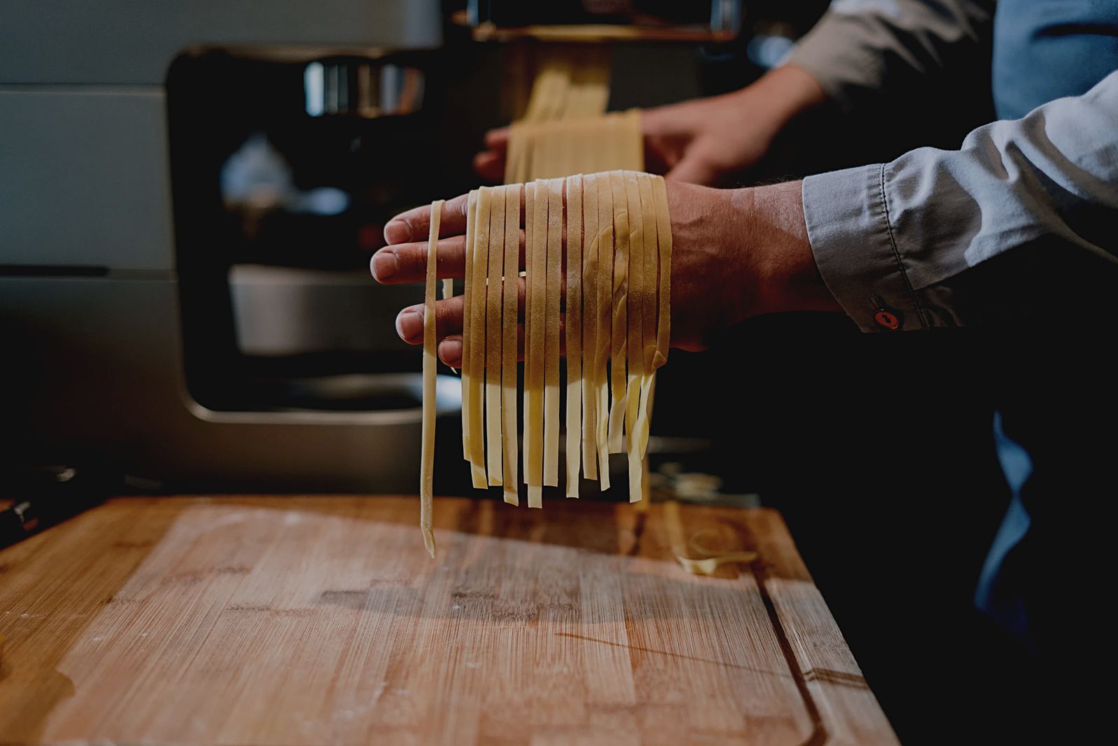 Close-up of Felix Kris's hands catching long strands of freshly cut pasta over the back of his hand, with a pasta machine and a floured wooden board